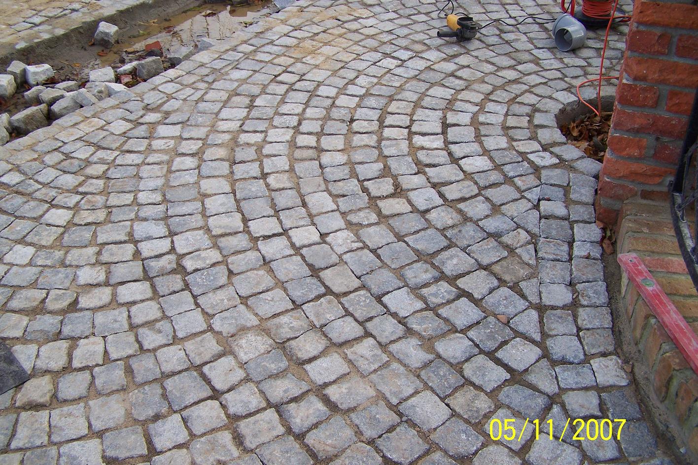 Układanie kostki brukowej z kamienia naturalnego w łuk, widoczne narzędzia pracy i poziomica przy murze.